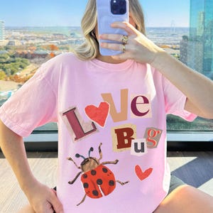 Pode incluir: Camiseta rosa com um gráfico de uma joaninha e o texto "LOVE BUGS" em letras coloridas recortadas.