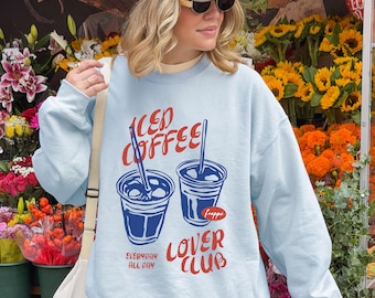 May include: A light blue sweatshirt with the text "ICED COFFEE LOVER CLUB" in red and white. The graphic features two iced coffee cups with straws. The person is wearing a beige cap and sunglasses.