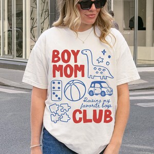White Comfort Colors boy mom club shirt featuring dinosaur toy car puzzle ball illustrations with raising my favorite boys slogan motherhood graphic tee gift for moms of sons ships without price tag ready for gifting