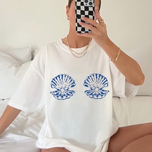 以下が含まれることがあります： 青い貝殻のデザインが施された白いTシャツ。デザインは、それぞれ中央に真珠がある2つの開いた貝殻が特徴です。ゆったりとしたオーバーサイズのフィット感のシャツです。