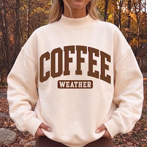 May include: A cream-colored long-sleeve sweatshirt with a mock neck collar. The front features the dark brown text COFFEE in a large collegiate font, with the word WEATHER printed in a smaller, rectangular brown box directly underneath.