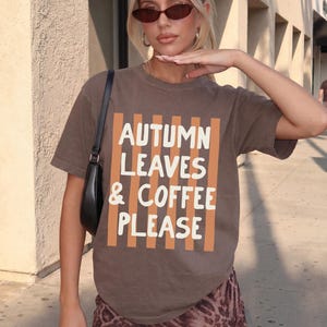 May include: A brown short-sleeved t-shirt featuring a graphic print of vertical orange stripes and the white text "AUTUMN LEAVES & COFFEE PLEASE" centered on the front. The shirt has a crew neckline and a relaxed fit, paired with a black shoulder bag.