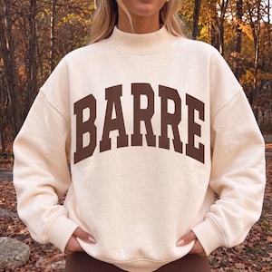 May include: A cream-colored long-sleeve sweatshirt with a mock neck collar. The front features the word BARRE printed in large, dark brown, block-style capital letters with a slight arch across the chest area.
