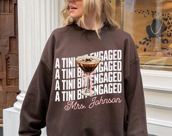 May include: A brown sweatshirt with the words "A Tini Bit Engaged" in white, along with a graphic of a martini and the name "Mrs. Johnson" in pink. The sweatshirt is a casual style.