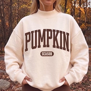 Op de afbeelding: Een crèmekleurig sweatshirt met lange mouwen en een opstaande kraag. De voorkant is voorzien van het woord PUMPKIN in grote, donkerbruine, gebogen blokletters, met het woord SEASON gedrukt in een kleiner donkerbruin ovaal onder de tekst.