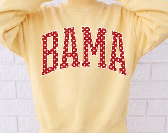 May include: A pale yellow long-sleeve sweatshirt featuring a mock neck and ribbed cuffs. The front displays the word BAMA in large, arched, red block letters with a white polka dot pattern and a thin white outline.