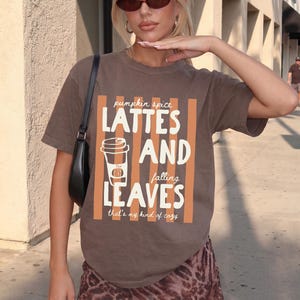 May include: A brown short-sleeved t-shirt featuring a graphic with vertical orange stripes and white text that reads: pumpkin spice LATTES AND falling LEAVES that's my kind of cozy, along with a line-art illustration of a coffee cup with a pumpkin.