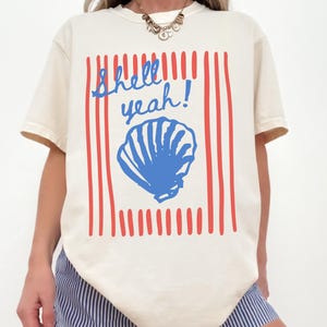 May include: Cream-colored t-shirt with a blue seashell graphic and the phrase "Shell yeah!" in blue script. The design includes red vertical stripes. The shirt is a casual, beach-themed apparel item.