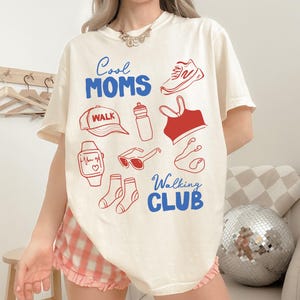 May include: Cream-colored t-shirt with the text "Cool Moms Walk" in blue and red. The shirt features red illustrations of walking-related items like a shoe, hat, water bottle, and socks. The shirt is a casual wear item.