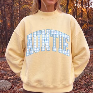 May include: A pale yellow long-sleeve sweatshirt with a mock neck collar. The front features the word AUNTIE printed in a large, arched collegiate font with a light blue and white gingham checkered pattern throughout the letters.