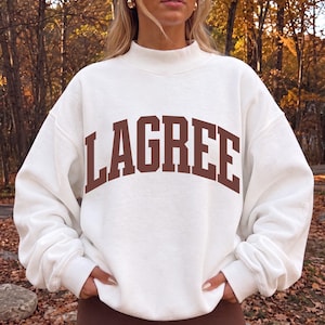 May include: An off-white crewneck sweatshirt featuring a mock neck collar and long sleeves. The word LAGREE is printed across the chest in large, arched, dark brown capital letters. The garment has a relaxed fit with ribbed cuffs.