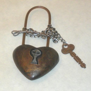 Puede incluir: Un candado en forma de corazón con una llave. El candado es de color bronce con una cadena plateada y una llave. La cerradura presenta un diseño alienígena. La llave también es de color bronce.