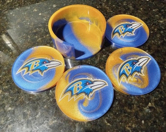 May include: Set of four blue and gold coasters with a Baltimore Ravens logo. The coasters are made of resin and have a glossy finish. The set also includes a matching coaster holder.