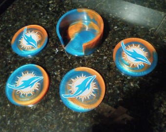 Puede incluir: Conjunto de seis posavasos azules y naranjas con un logotipo de delfín. Los posavasos están hechos de resina y tienen un acabado brillante. El conjunto incluye un portavasos a juego.