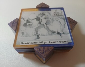 Peut inclure: Un dessous de verre décoratif avec une image en noir et blanc d'un joueur de football américain courant avec le ballon. Le dessous de verre a une bordure bleue et dorée, avec le texte "Jacoby Jones 108 yd. kickoff return". Il a quatre coins triangulaires.