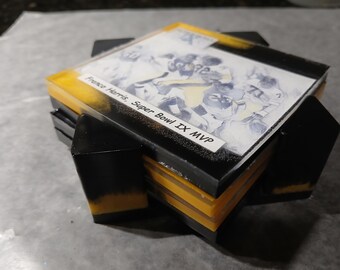 May include: A stack of square coasters with a football theme. The top coaster features a black border, a yellow centre, and a picture of a football team. Text on the coaster reads "Franco Harris, Super Bowl IX MVP". The coasters are black and yellow.
