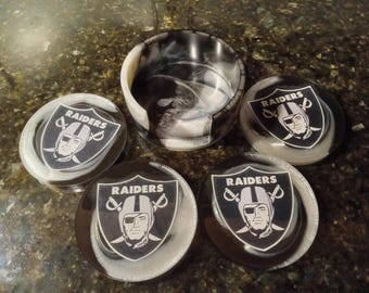 Puede incluir: Conjunto de cuatro posavasos redondos en blanco y negro con un logotipo de Raiders plateado y la imagen de un jugador. Los posavasos tienen un patrón de remolino y están hechos de resina.