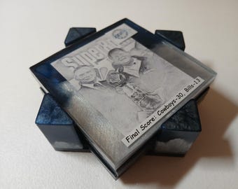 May include: A square coaster with a black and white image of two men holding a trophy, with the word "Super Bowl" above them. The coaster is set in a clear resin with blue accents and rests on a dark blue base. The text "Final Score: Cowboys-30, Bills-13" is visible.