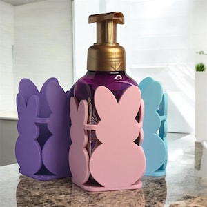 May include: Three soap dispensers in bunny-shaped holders. The holders are purple, pink, and blue. The soap dispensers are clear with gold tops.