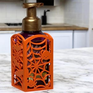 May include: Orange Halloween-themed soap dispenser holder with a spider web, bats, and smiling spiders. The holder is designed to fit around a soap bottle, adding a festive touch to any bathroom or kitchen during the spooky season.