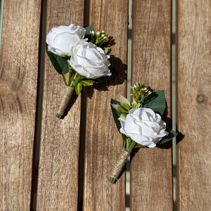 May include: Two white rose boutonnieres with green leaves and small buds, wrapped with twine. The floral arrangements are set on a wooden surface, perfect for weddings or special events.