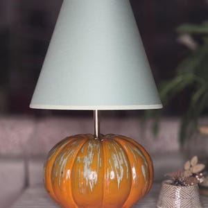 Può includere: Una lampada da tavolo con base a forma di zucca, caratterizzata da una smaltatura arancione lucida con accenti bianchi colanti. La lampada presenta uno stelo metallico sottile e un paralume conico di colore azzurro uniforme e diffuso.