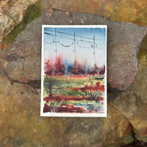 May include: A rectangular watercolor painting on paper depicting a landscape with a blue sky, utility poles with power lines, and a field featuring patches of green grass and deep red foliage. The artwork has a white border and a textured surface.