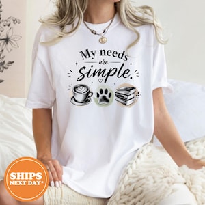 May include: A white short-sleeve t-shirt featuring the black text My needs are simple. The design includes a black and white illustration of a coffee cup, a paw print, and a stack of three books, all arranged horizontally across the chest.