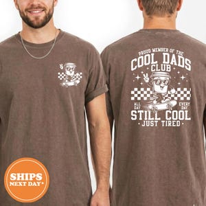 May include: A brown short-sleeve t-shirt featuring a white graphic of a coffee cup on a skateboard. The front chest displays a small version, while the back features a larger graphic with the text "PROUD MEMBER OF THE COOL DADS CLUB STILL COOL JUST TIRED".