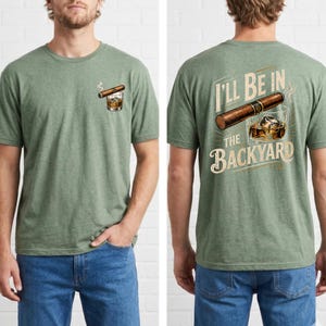 May include: A heather green short-sleeve t-shirt featuring a small graphic on the front left chest of a cigar and a glass of whiskey. The back displays a large graphic of a cigar and a glass of whiskey with the text I'LL BE IN THE BACKYARD.