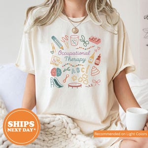 May include: A cream-colored t-shirt featuring a colorful graphic print with the text Occupational Therapy. The design includes illustrations of a pencil, scissors, glue, a brain, a slide, a trampoline, puzzle pieces, and the letters ABC.