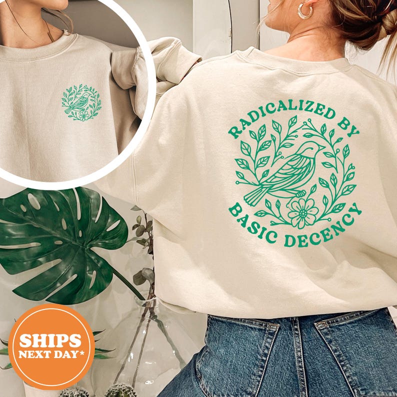 a person wearing a white sweatshirt with a green graphic design on the back. The design includes a bird and leaves, and the text "RADICALLY BY BASIC DECENCY" is printed on the sweatshirt.