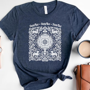 A navy blue t-shirt with a white graphic design featuring a sunburst and religious symbols, along with the text "Sanctus + Sanctus + Sanctus" printed on it. The t-shirt is placed on a white surface, with a cup, a handbag, and a watch nearby.