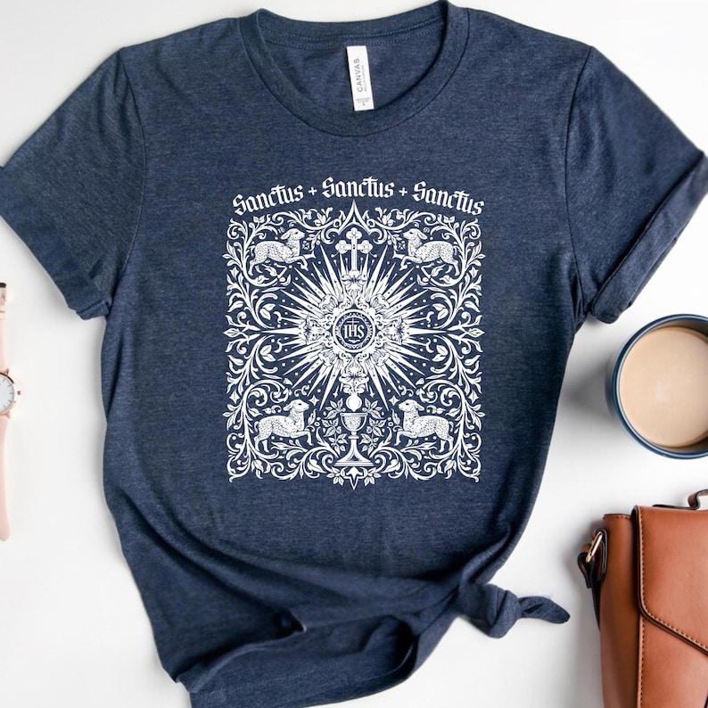 A navy blue t-shirt with a white graphic design featuring a sunburst and religious symbols, along with the text "Sanctus + Sanctus + Sanctus" printed on it. The t-shirt is placed on a white surface, with a cup, a handbag, and a watch nearby.