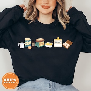 May include: A black long-sleeve sweatshirt featuring a graphic print of baking ingredients including a measuring cup, a milk carton, a vanilla bottle, an egg, a bag labeled flour, a bowl of butter, a sugar jar, and a carton of eggs.