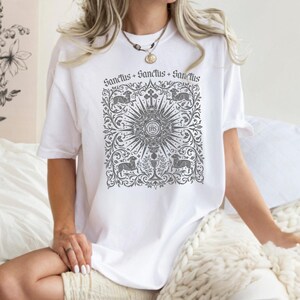A woman wearing a white t-shirt with a graphic design printed on it, sitting on a bed with a white blanket.