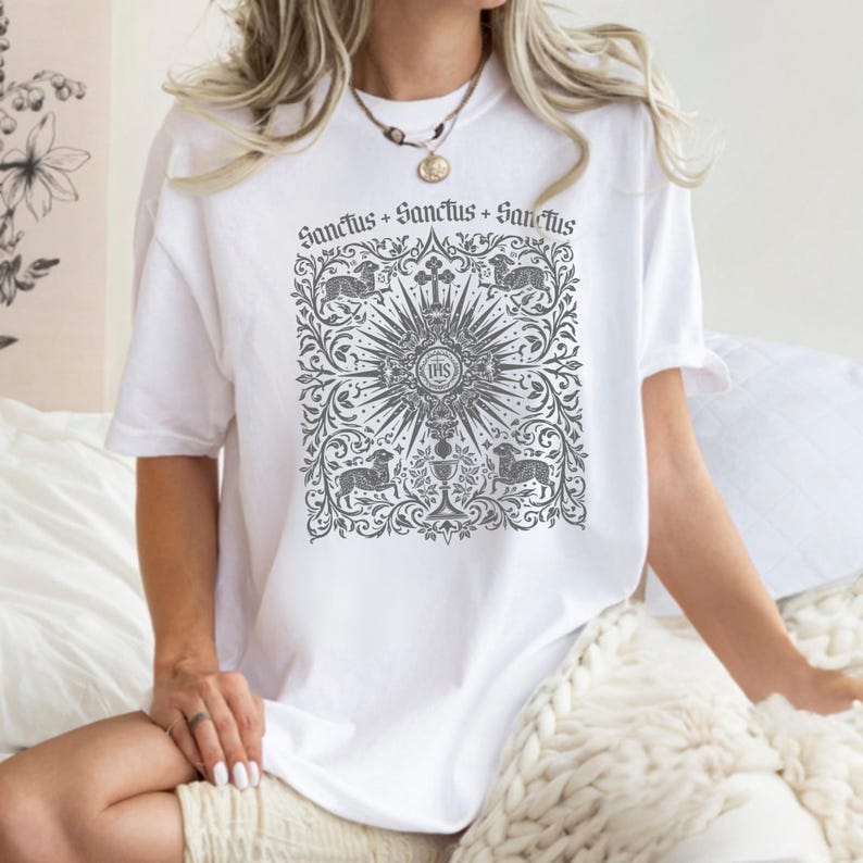A woman wearing a white t-shirt with a graphic design printed on it, sitting on a bed with a white blanket.