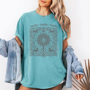 A woman wearing a turquoise t-shirt with a graphic design and a denim jacket, standing against a white background.