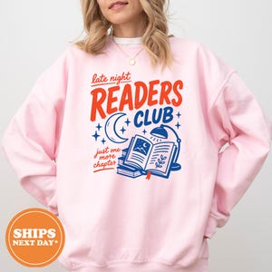 May include: Pink sweatshirt with the text "late night READERS CLUB" in red and blue. The design includes a crescent moon, stars, an open book, and a desk lamp. The phrase "just one more chapter" is also included.