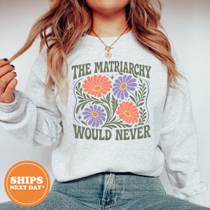 May include: A light gray crewneck sweatshirt featuring a graphic print of orange and purple daisies with green leaves. The text reads THE MATRIARCHY WOULD NEVER in a dark green font. The design is centered on the chest of the long-sleeved garment.