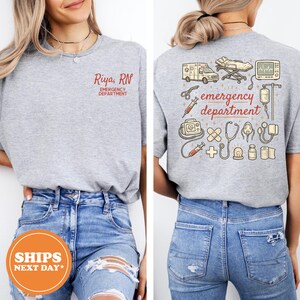 May include: A heather gray t-shirt featuring a small red graphic on the front that reads Riya, RN EMERGENCY DEPARTMENT. The back displays a large graphic with medical icons and the text emergency department in red, including an ambulance.