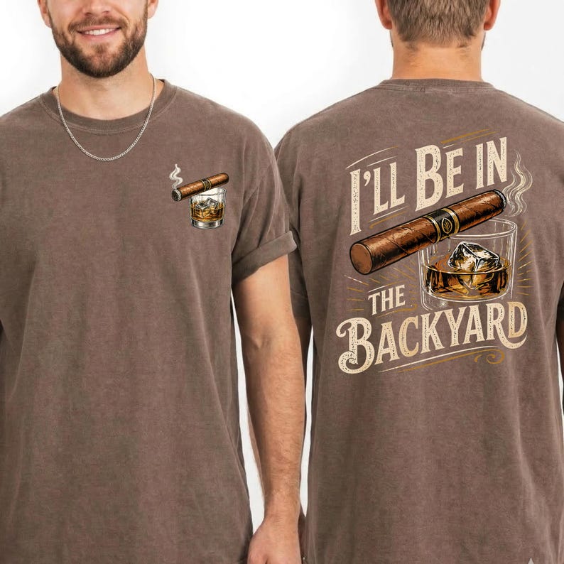 May include: A brown short-sleeve t-shirt featuring a small graphic on the front left chest of a cigar and a glass of whiskey with ice. The back of the shirt displays a large cigar, a glass of whiskey with ice, and the text I'LL BE IN THE BACKYARD.