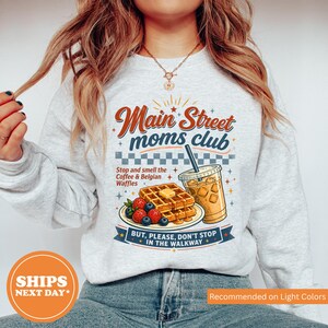 May include: Light gray sweatshirt with the text "Main Street moms club" in a retro design. The graphic includes waffles, berries, iced coffee, and the text "Stop and smell the Coffee & Belgian Waffles".