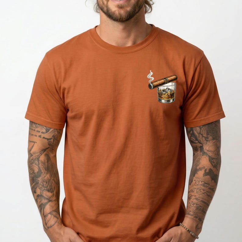 May include: A rust-colored short-sleeve t-shirt featuring a graphic on the left chest area. The graphic depicts a brown cigar resting across the rim of a clear glass filled with an amber-colored liquid and ice cubes, with a small wisp of smoke.