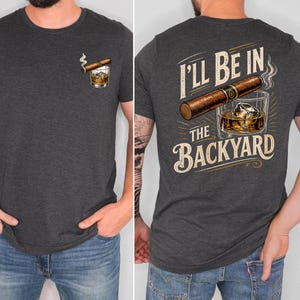 May include: A dark charcoal gray t-shirt featuring a small graphic on the front left chest of a cigar resting on a glass of whiskey with ice. The back of the shirt displays a large cigar, a glass of whiskey with ice, and the text I'LL BE IN THE BACKYARD.