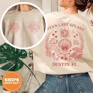 May include: Beige sweatshirt with red seashell graphics and text. The front features two seashells and "RIYA'S BACHELORETTE". The back has a mermaid, disco ball, and text that reads "RIYA'S LAST SPLASH, Est. 2026, DESTIN, FL".