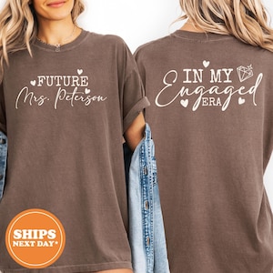 May include: A brown short-sleeved t-shirt featuring white text. The front displays "FUTURE Mrs. Peterson" with small hearts. The back shows "IN MY Engaged ERA" with a diamond icon and hearts. The shirt is shown from both front and back views.