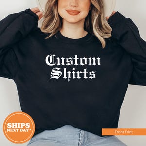 May include: Black sweatshirt with the words "Custom Shirts" in a white gothic font. The image also includes a round orange sticker that says "Ships Next Day" and an orange rectangle that says "Front Print".
