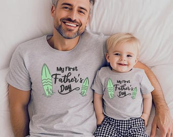 May include: Matching grey t-shirts with the text "My First Father's Day" and a surfboard graphic. One shirt is worn by an adult, the other by a child. The child is wearing black and white checkered trousers.