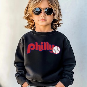 Könnte beinhalten: Ein schwarzer Pullover mit dem Wort "philly" in Rot und einer Baseballgrafik. Der Pullover wird von einem kleinen Kind getragen, das eine braune Sonnenbrille trägt.